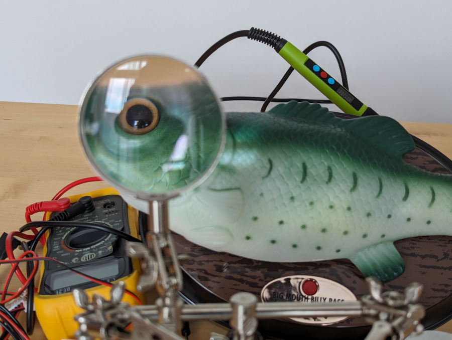 Billy Bass and a pile of electronics gear. The magnifying glass of the "helping hand" is magnifying the eye of the fish.